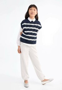 Temno modra pletena brezrokavnica z belimi vodoravnimi črtami, v kombinaciji z belo srajco s ovratnikom in svetlo sivo zoženimi hlačami. Srebrni balerinki.