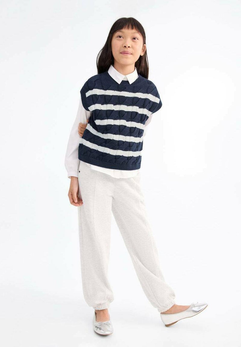 Temno modra pletena brezrokavnica z belimi vodoravnimi črtami, v kombinaciji z belo srajco s ovratnikom in svetlo sivo zoženimi hlačami. Srebrni balerinki.