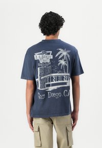 Mörkblå bomull t-shirt med en grafisk design av ett grillskylte, palmträd och texten "San Diego, CA" tryckt i vitt på baksidan.