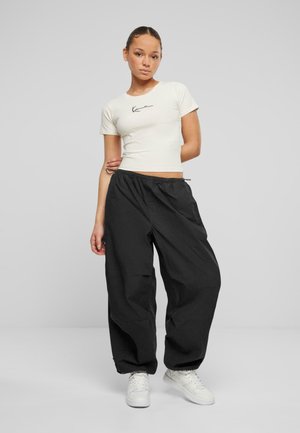 T-shirt ajusté beige clair avec un logo noir, associé à un pantalon de jogging noir ample et des baskets blanches, doté d'une taille élastique avec cordons de serrage.