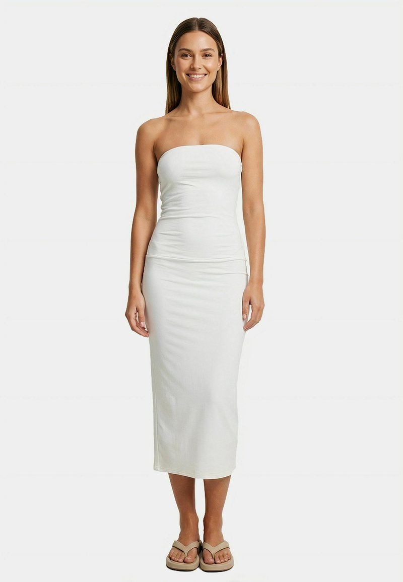 Femme souriante aux longs cheveux portant une robe midi blanche sans bretelles et des tongs beige, debout devant un fond clair uni.