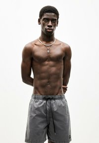 Männliche graue Badeshorts mit Kordelzug, aus leichtem Material gefertigt. Das Model hat einen durchtrainierten Körper und trägt mehrere Halsketten übereinander.