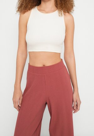 Femme aux cheveux bouclés portant un crop top blanc sans manches et un pantalon ample taille haute couleur rouille sur un fond uni.