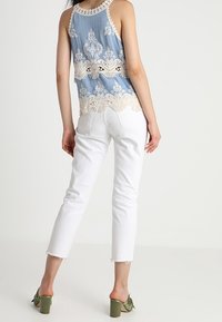 Jasnoniebieski top na ramiączkach z kremowymi koronkowymi akcentami, białe krótkie dżinsy i zielone sandały na słupku. Widok z tyłu ukazuje koronkowe detale i dopasowanie.