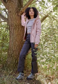 Veste rose clair sur un haut gris ajusté, associée à un pantalon foncé et des bottes de randonnée grises avec des lacets bleus. Entourée d'arbres et de verdure.