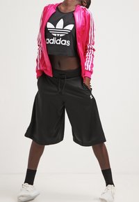 Rosa satin-jacka med vita ränder, svart crop top med vit adidas-logga, svarta shorts och vita sportskor.