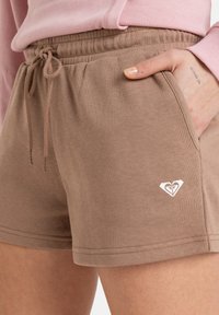 Shorts en coton marron avec une taille élastique dotée d'un cordon de serrage et d'un petit logo blanc sur le bas à gauche.