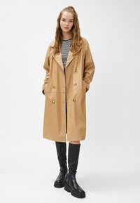 Beige syntetläder trench coat med dubbelknäppt design, stora slag, sidofickor och avslutat med svarta knähöga stövlar.