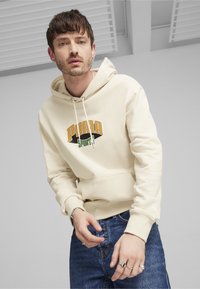 Felpa crema con cappuccio caratterizzata da un audace logo "PUMA" in arancione e verde. Materiale in pile testurizzato con tasca a marsupio e cappuccio con lacci.