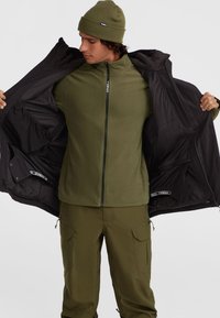 Veste noire sur un haut en polaire vert olive et un pantalon assorti. La veste présente une texture lisse avec des zips prononcés et des poches intérieures.