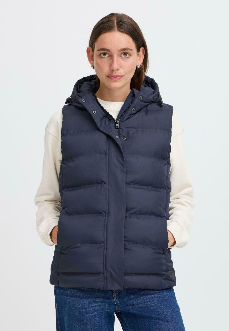 Une jeune femme aux longs cheveux porte un gilet matelassé bleu marine sur un pull léger, se tenant contre un fond uni, les mains dans les poches.
