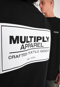 Musta puuvillapaita, jossa on suuri valkoinen etiketti takana mustalla tekstillä, jossa lukee "MULTIPLY APPAREL" ja "CRAFTED TEXTILE GOODS."