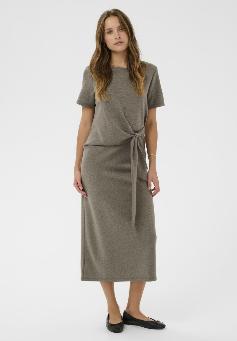 Knee-dolžine olivno zelena obleka iz bombažne mešanice s kratkimi rokavi, z zvitim vozlom na pasu in ravno silhueto. Črne ravne čevlje.