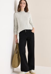 Jasnoszary sweter z warkoczowym splotem i wysokim kołnierzem, zestawiony z czarnymi spodniami typu wide-leg. Model trzyma khaki torbę. Białe sneakersy z niebieskimi akcentami.