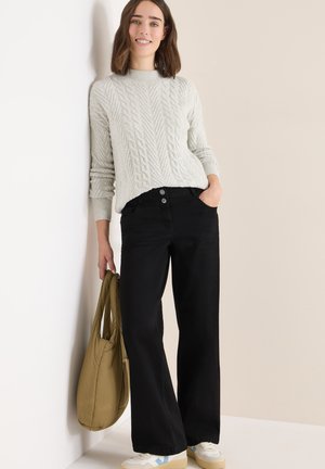 Suéter de punto grueso gris claro con cuello alto, combinado con pantalones vaqueros anchos negros. La modelo lleva un bolso caqui. Zapatillas blancas con detalles en azul.