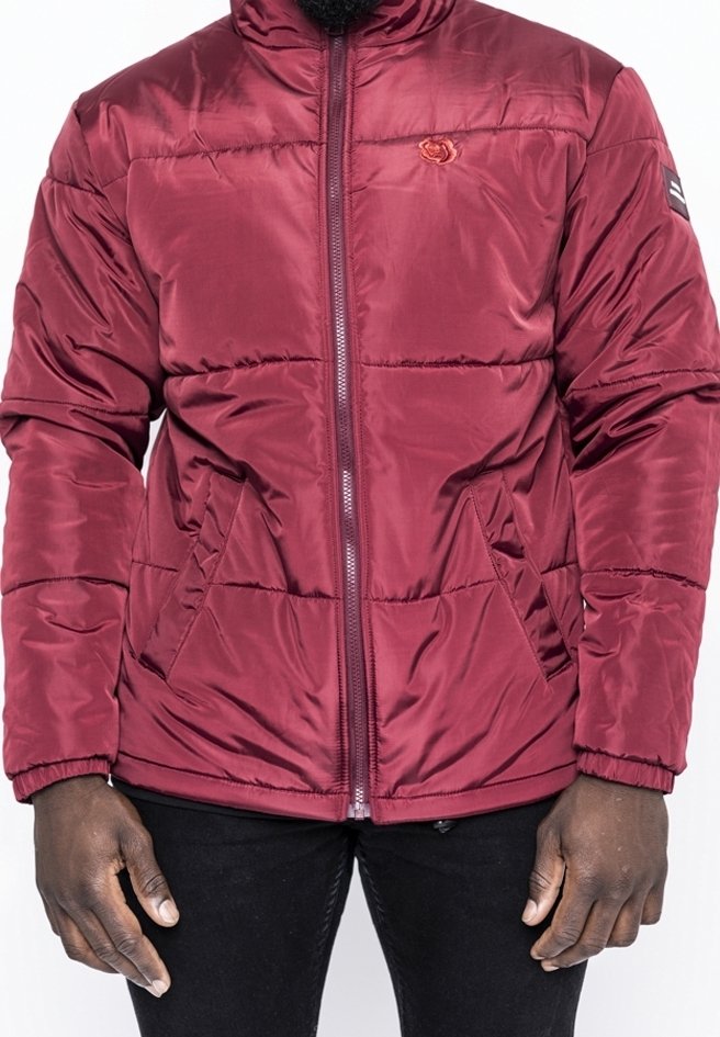 Mann trägt eine kastanienbraune gesteppte Jacke mit Frontreißverschluss und zwei Seitentaschen über schwarzer Hose, steht mit entspannten Armen.