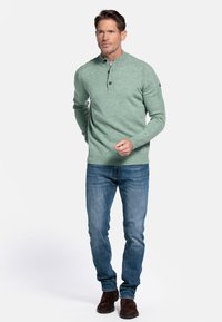 Lichtgroene, gestructureerde gebreide trui met een henleyhals, drie knopen en ribbelboorden, gecombineerd met blauwe denim jeans en bruine schoenen.