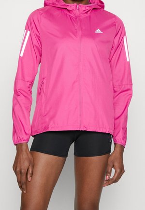 Personne portant une veste à capuche Adidas rose vif avec fermeture éclair à l’avant et un short de sport noir sur un fond uni.