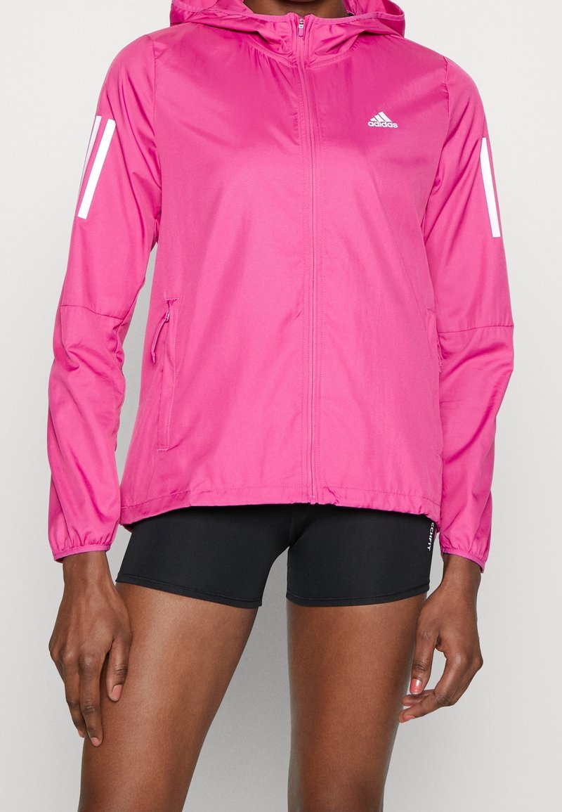 Personne portant une veste à capuche Adidas rose vif avec fermeture éclair à l’avant et un short de sport noir sur un fond uni.