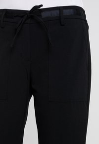 Pantalon noir à taille élastique avec cordon de serrage, doté de deux poches avant et d'une texture lisse. Le tissu a une finition mate.
