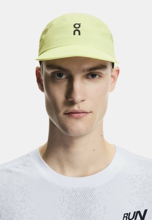 Mladý muž nosící světle žlutou čepici s černým logem a bílým sportovním tričkem s nápisem "RUN" na přední straně.