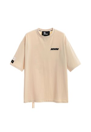 Camiseta beige de manga corta oversize con cuello redondo, logo negro "MWM" en el pecho, etiqueta dentro del cuello y pequeña etiqueta en la manga izquierda.