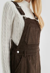 Braune Corduroy-Overalls mit verstellbaren Trägern und einer Vordertasche, kombiniert mit einem weißen, strukturierten Strickpullover, der Textur und Kontrast zur Geltung bringt.