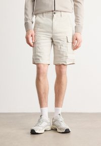 Beige cargo shorts med front- og sidelommer, oppbrettede cuffs og knappefeste. Brukt med hvite ankelsokker og grå sneakers.