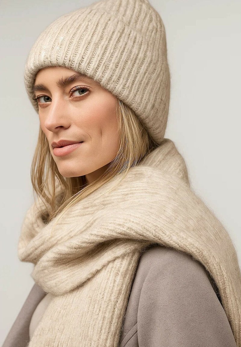 Beige geribbelde gebreide muts met pailletten, gecombineerd met een zachte, gedrapeerde sjaal in hetzelfde materiaal. Fijne textuur en warme uitstraling.