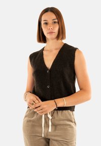 Gilet a maglina marrone senza maniche con scollatura a V profonda e due bottoni sul davanti. Indossato sopra pantaloni in corduroy chiari.