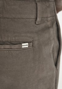 Brune bomuldsbukser med en baglomme, hvor der er syet et mærke med teksten "Resteröds." Teksturen er glat med et subtilt vævet mønster.