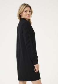 Schwarzes Kleid aus weichem Stoff, mit plissierten langen Ärmeln und lockerer Passform. Der Ausschnitt ist schlicht, und der Saum endet leicht über dem Knie.