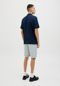 Marineblaues Polohemd mit kurzen Ärmeln, kombiniert mit hellgrauen, hochgekrempelten Shorts. Schwarze Schuhe und weiße Socken runden das Outfit ab. Schlichtes Design.