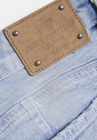 Φωτεινό μπλε τζιν με δερμάτινη ταμπέλα σε καφέ χρώμα που φέρει την επιγραφή "Juicy Couture Jeans", διακοσμημένο με ασημένια καρφιά και προσεκτή ραφή.