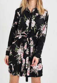 Svart långärmad klänning med rosa och vit blommig och fågeltryck, knäppt framtill och med ett matchande bälte av samma tyg knytet i midjan.