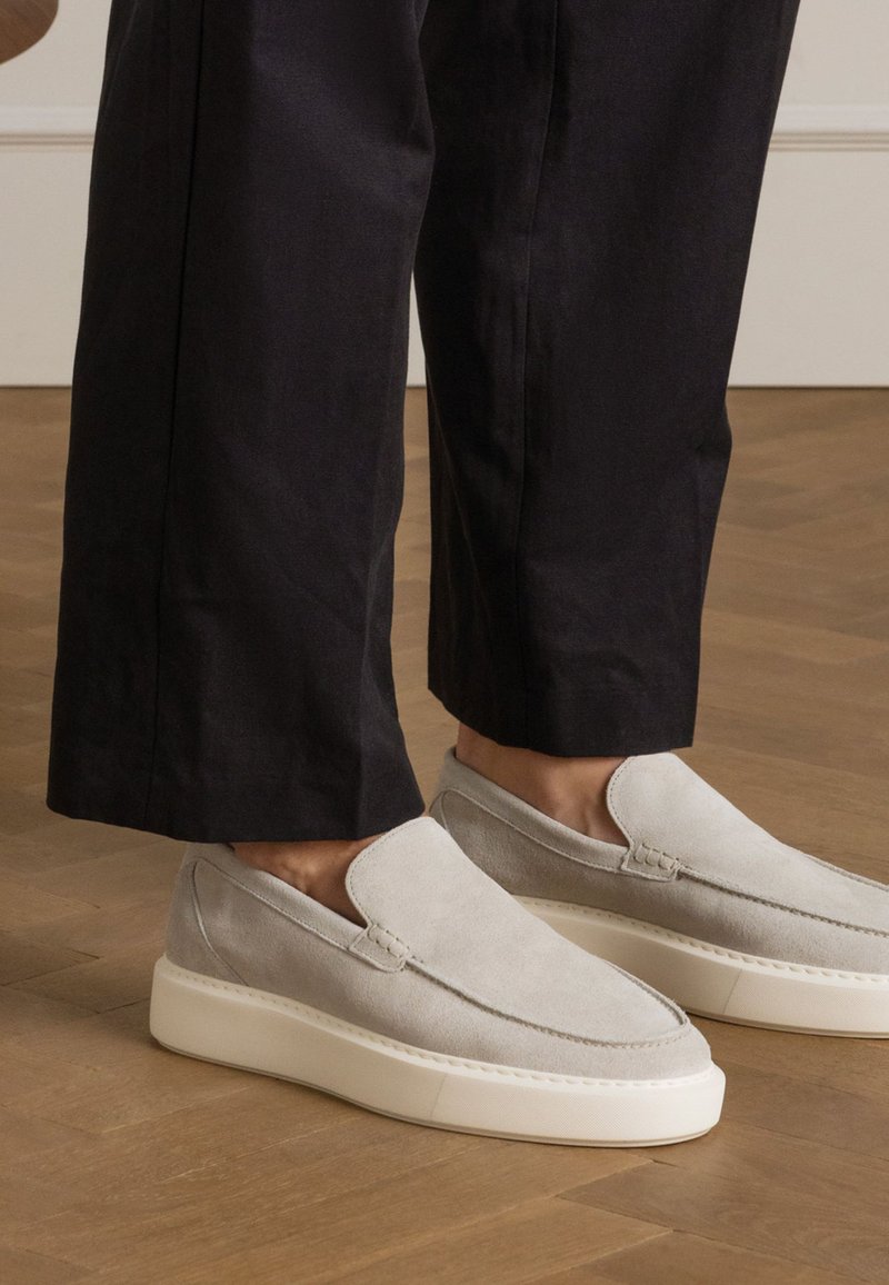 Light gray suede slip-on shoes with a rounded toe and white sole, paired with black wide-leg trousers on a wooden floor.