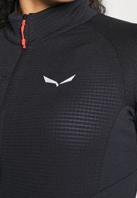 Svart idrottsjacka med hög krage, med ett texturerat mönster på tyget. Märkbar orange dragkedja och en silverlogotyp som accent.