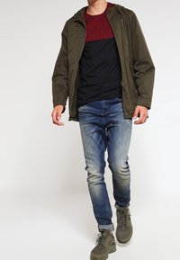 Olivgrön jacka, skjorta med röd och svart mönstring, blå jeans och grå sneakers. Jackan har huva och framdragkedja.