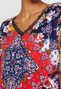 Punainen kukkakuosiset pusero, jossa on laivastonsininen yksityiskohta, V-kaula-aukko ja hieno musta pitsireunus kaula-aukolla.