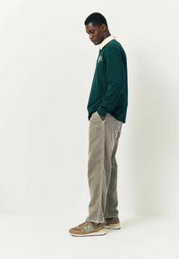 Grünes Langarm-Polohemd mit weißem Kragen, kombiniert mit beigen Cordhosen. Getragen mit braunen und grauen Sneakern mit grünem Logo.