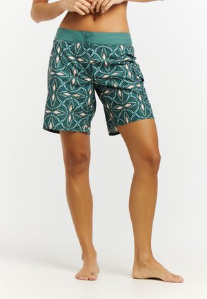 Frau trägt grüne, gemusterte Shorts in Kniehöhe mit zwei vorderen Knöpfen und einer Cargotasche, steht barfuß vor weißem Hintergrund.