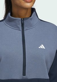 Grijze sweatshirt met hoge kraag, halve rits en marineblauwe accenten. Bevat een wit logo op de borst en een zachte, gestructureerde stof.