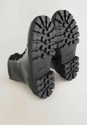 Juodi odiniai bateliai su raišteliais matomi iš apačios, rodančiais storas, tvirtas padangas su "NEXT" ženklu kiekvienoje padangoje.