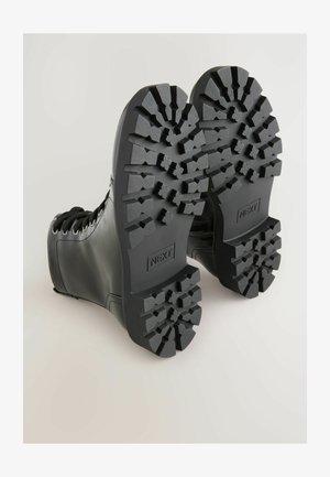 Mustad mustad nahast nööriga saapad vaadatuna altpoolt, näidates paksu robustset tallamustrit, millel on iga talda peal "NEXT" bränding.