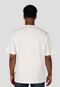 Bílý krátký rukáv tričko zobrazuje zadní pohled s přímým švem uprostřed a obdélníkovou visačkou blízko výstřihu. Měkká tkanina.