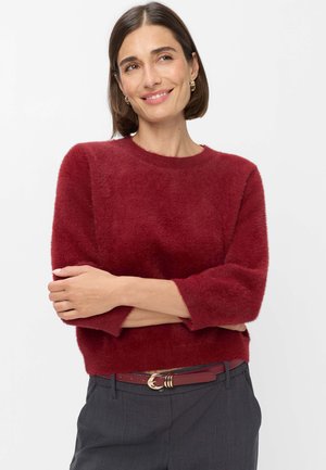 Vrouw met kort bruin haar die gouden ronde oorbellen draagt, een rode pluizige trui, donkergrijze broek en een rode riem, staand met gekruiste armen.