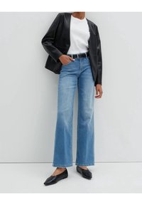 Person som bär ljusblå vida jeans, vit t-shirt, svart läderjacka, svart bälte och svarta spetsiga ballerinas, stående mot en vit vägg.