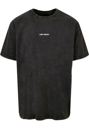 Mand iført en falmet sort t-shirt med teksten "LOST YOUTH™" på brystet, hvide bukser, sølvkædehalskæde og synlige armtatoveringer.