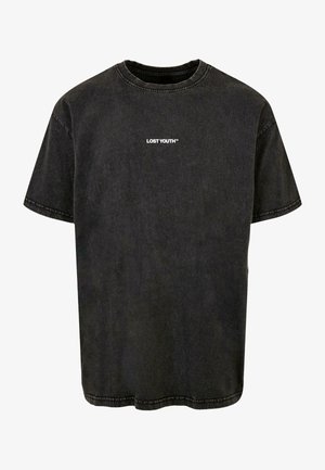 Mężczyzna w wyblakłym czarnym T-shircie z napisem "LOST YOUTH™" na piersi, białych spodniach, srebrnym łańcuszku oraz widocznymi tatuażami na ramionach.