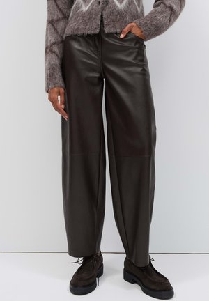 Persona che indossa pantaloni neri in pelle a gamba larga, scarpe stringate in camoscio marrone e un maglione grigio lavorato a maglia con motivi, con le mani nelle tasche.